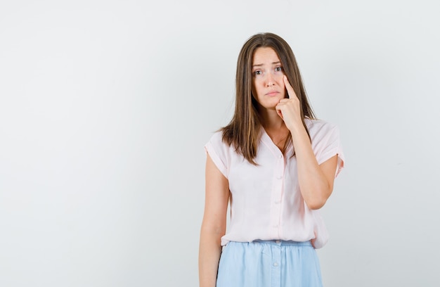 Jeune fille en t-shirt, jupe pointant le doigt sur son œil et à la malheureuse, vue de face.