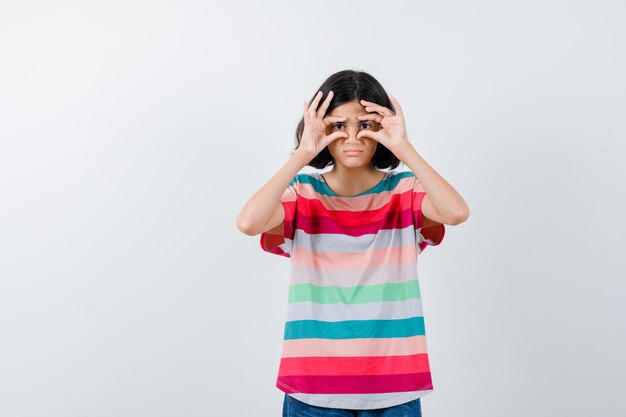 Jeune fille en t-shirt rayé coloré montrant le geste des jumelles et l'air mignon, vue de face.