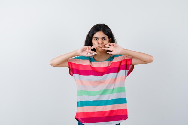 Jeune fille en t-shirt à rayures colorées mettant l'index sur les joues et l'air sérieux, vue de face.