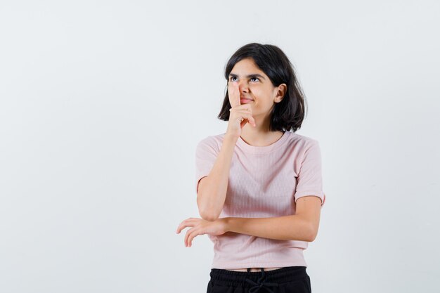 Jeune fille en t-shirt rose et pantalon noir debout en pensant poser et mettre l'index sur le nez et à la recherche de bonheur