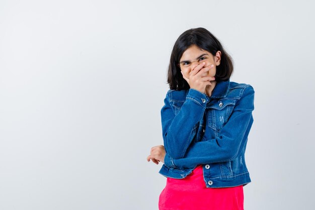 Jeune fille en t-shirt rouge et veste en jean couvrant la bouche avec la main, riant et ayant l'air heureux, vue de face.