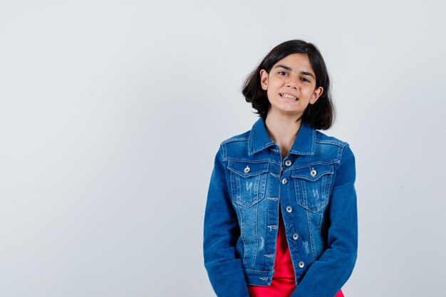 Jeune fille en t-shirt rouge et veste en jean debout tout droit et se présentant à la caméra et l'air heureux, vue de face.