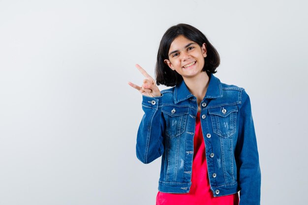 Jeune fille en t-shirt rouge et veste en jean montrant le geste rock n roll et l'air mignon, vue de face.