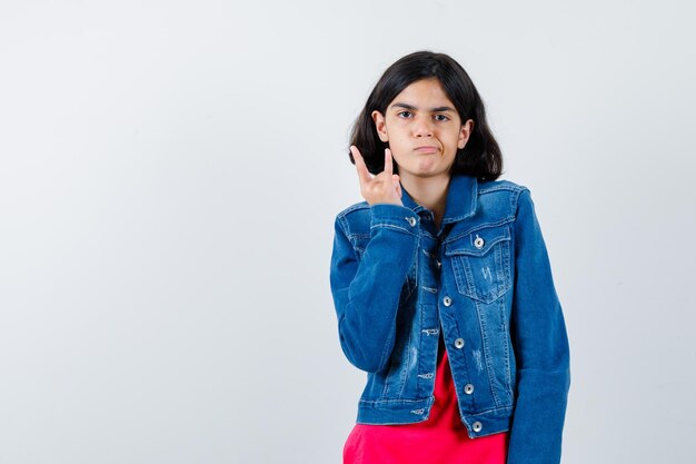 Jeune fille en t-shirt rouge et veste en jean montrant un geste rock n roll et l'air sérieux, vue de face.