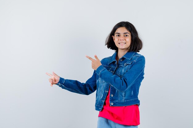 Jeune fille en t-shirt rouge et veste en jean pointant vers la gauche avec l'index et l'air heureux, vue de face.