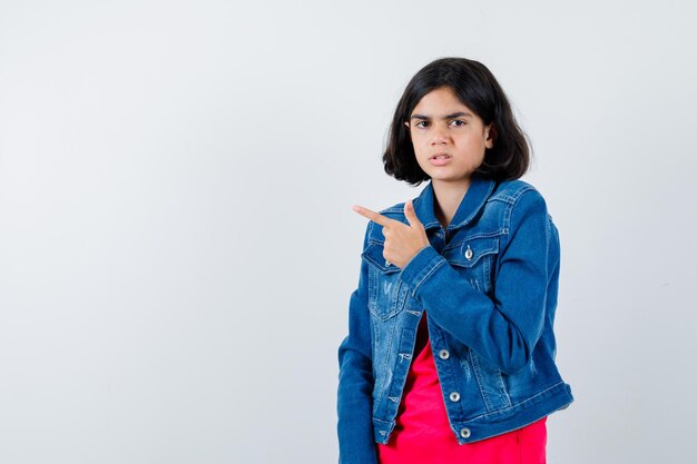 Jeune fille en t-shirt rouge et veste en jean pointant vers la gauche avec l'index et l'air sérieux, vue de face.