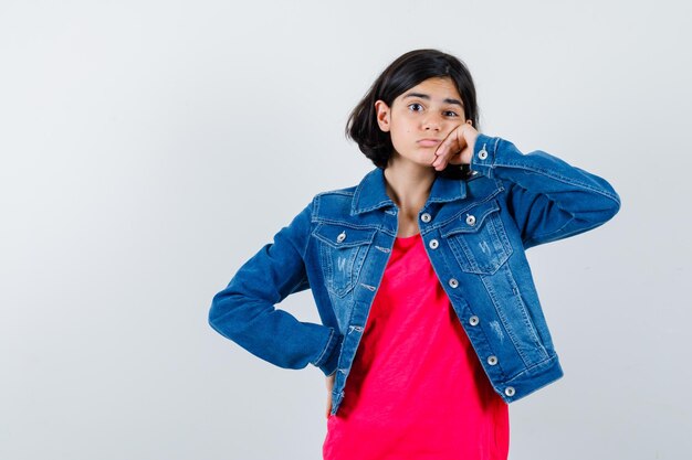 Jeune fille en t-shirt rouge et veste en jean s'appuyant sur la joue de la paume, tenant la main sur la taille et l'air pensif