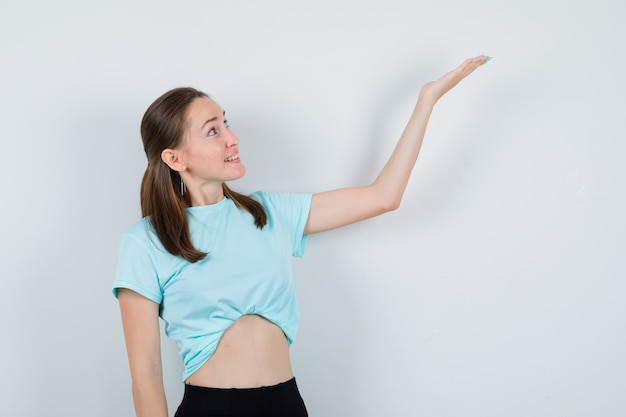 Jeune fille en t-shirt turquoise, pantalon écartant le bras et l'air curieux, vue de face.