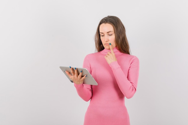 Jeune fille avec tablette pense en tenant l'index sur les lèvres sur fond blanc