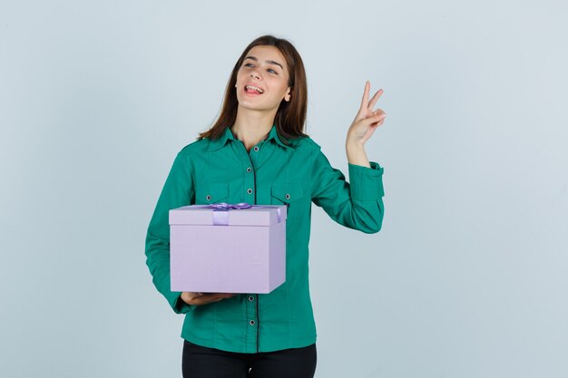 Jeune fille tenant une boîte-cadeau, montrant le geste de paix en chemisier vert, pantalon noir et à la joyeuse, vue de face.