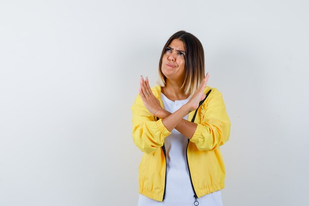 Jeune fille tenant deux bras croisés, ne faisant aucun signe, courbes des lèvres en t-shirt blanc, veste jaune et à la vue inquiète, de face.