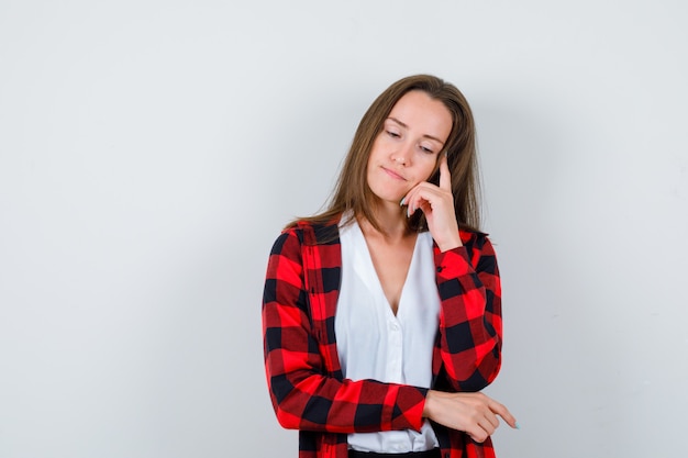 Jeune fille tenant le doigt sur les tempes en chemise à carreaux, chemisier et regardant réfléchie, vue de face.