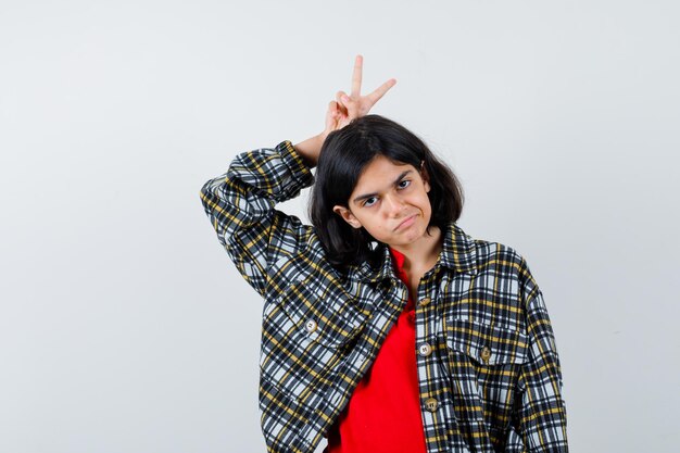 Jeune fille tenant les doigts au-dessus de la tête comme geste de paix en chemise à carreaux et t-shirt rouge et l'air sérieux, vue de face.