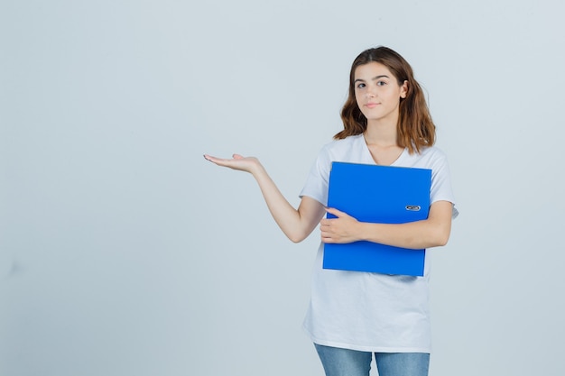 Jeune fille tenant le dossier, montrant un geste de bienvenue en t-shirt blanc et regardant confiant, vue de face.