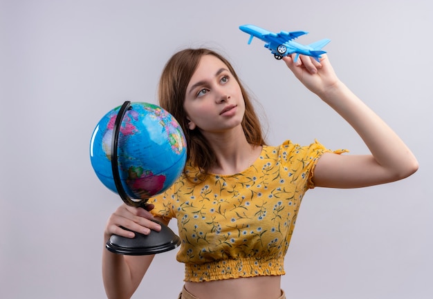 Photo gratuite jeune fille tenant un globe et un avion modèle et regardant un avion modèle sur un mur blanc isolé