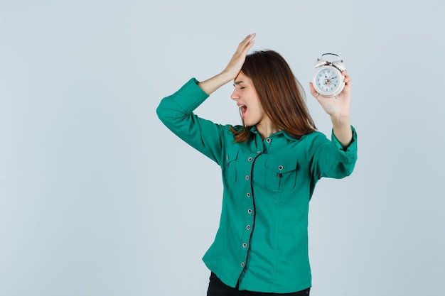 Jeune fille tenant une horloge, mettant la main sur la tête en chemisier vert, pantalon noir et regardant harcelé, vue de face.