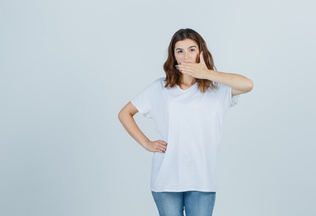 Jeune fille tenant la main sur la bouche en t-shirt blanc et à la surprise, vue de face.