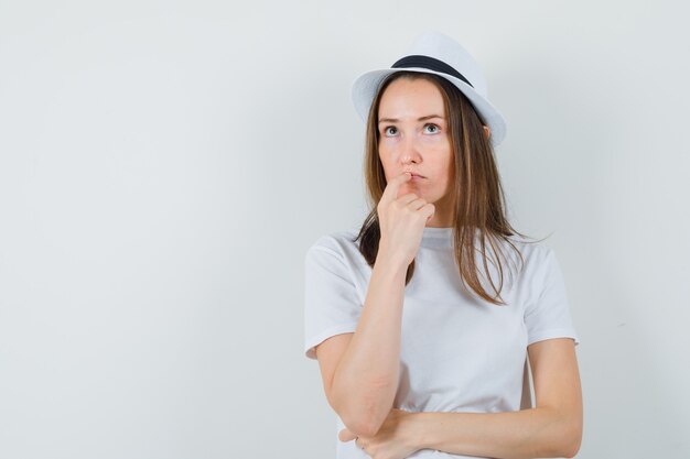 Jeune fille tenant la main sur le menton en t-shirt blanc, chapeau et à la réflexion. vue de face.