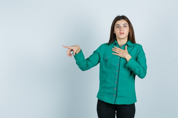 Jeune fille tenant la main sur la poitrine, pointant vers la gauche avec l'index en chemisier vert, pantalon noir et à la choqué. vue de face.