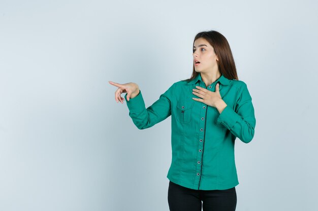 Jeune fille tenant la main sur la poitrine, pointant vers la gauche avec l'index en chemisier vert, pantalon noir et à la surprise, vue de face.