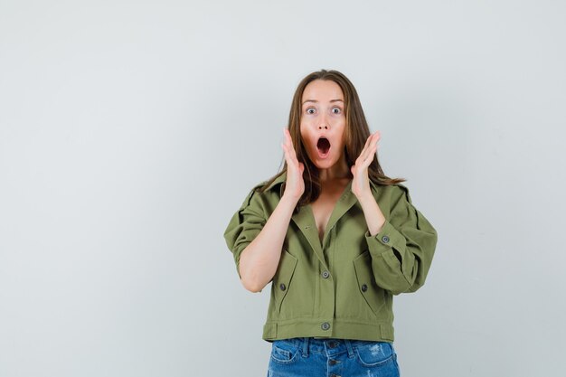 Jeune fille tenant la main près de la bouche ouverte en veste, short et à la vue surprise, de face.