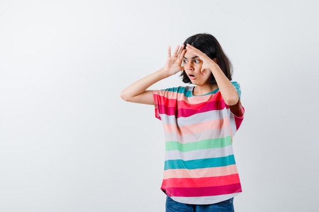 Jeune fille tenant la main près du visage en t-shirt rayé coloré et l'air surpris, vue de face.