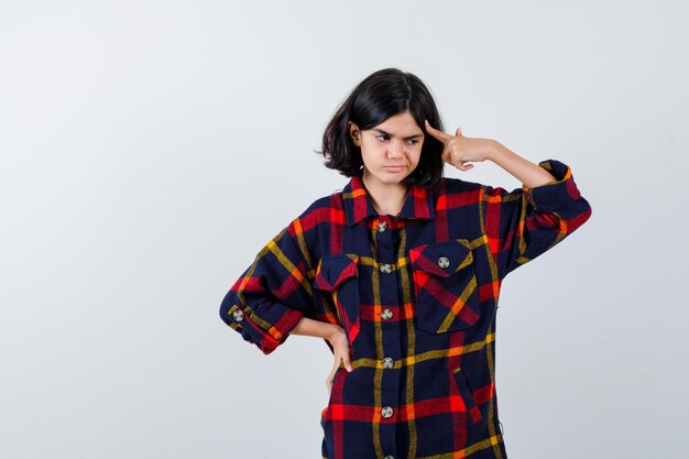 Jeune fille tenant la main sur la taille, mettant l'index sur la tempe en chemise à carreaux et l'air pensif, vue de face.