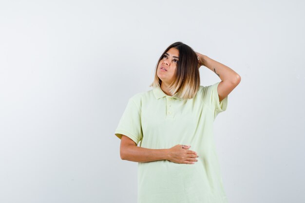 Jeune fille tenant une main sur la tête, une autre main sur le ventre, pensant à quelque chose en t-shirt et regardant pensif, vue de face.