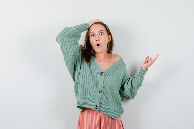 Jeune fille tenant une main sur la tête, pointant vers la droite avec l'index en tricot, jupe et à la surprise. vue de face.