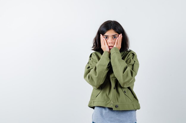 Jeune fille tenant les mains sur les joues en pull gris, veste kaki, pantalon en jean et l'air surpris. vue de face.