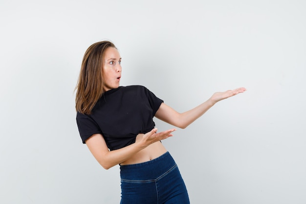 Jeune fille tenant des paumes pour montrer quelque chose de terrible en chemisier noir, pantalon et à la peur