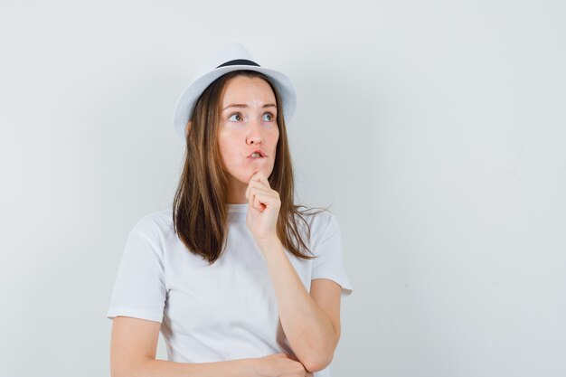 Jeune fille tenant son menton en t-shirt blanc, chapeau et à la vue de face, hésitante.