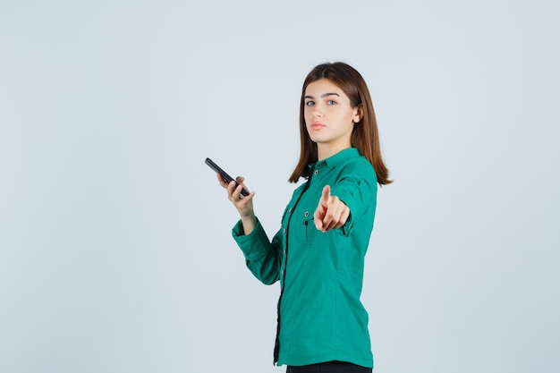 Jeune fille tenant le téléphone dans une main, pointant la caméra en chemisier vert, pantalon noir et à la recherche de sérieux. vue de face.