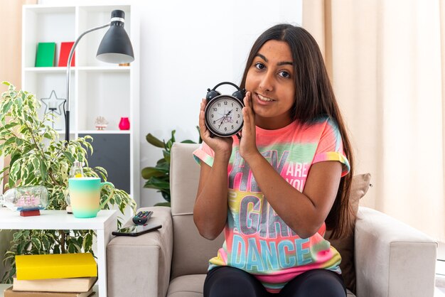 Jeune fille en tenue décontractée tenant un réveil à l'air heureux et positif souriant joyeusement assis sur une chaise dans un salon lumineux