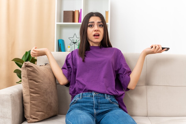 Jeune fille en tenue décontractée tenant la télécommande de la télévision à la confusion écartant les bras sur les côtés assis sur un canapé dans un salon lumineux