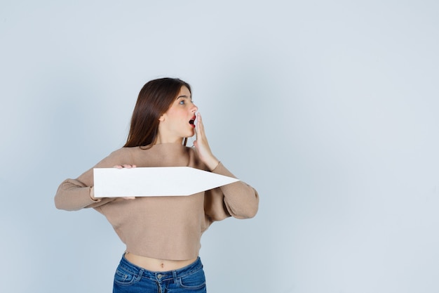 Jeune Fille En Tricot Beige, Jeans Tenant Un Bâton De Papier, Regardant Le Côté Droit, Couvrant La Bouche Avec La Main Et L'air Surpris, Vue De Face.