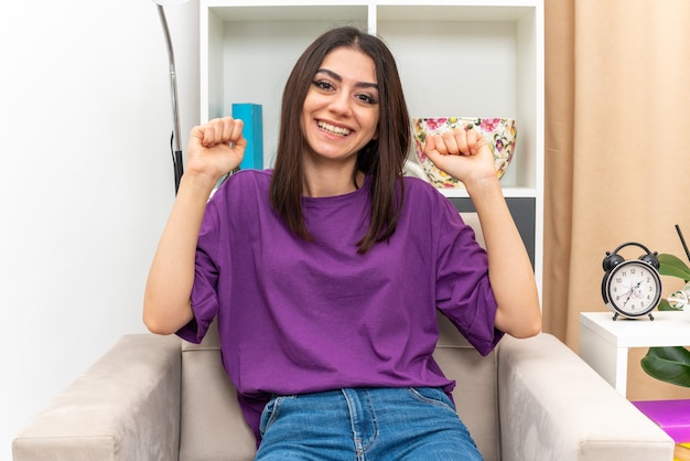 Jeune fille en vêtements décontractés, heureuse et excitée, serrant les poings, assise sur une chaise dans un salon lumineux