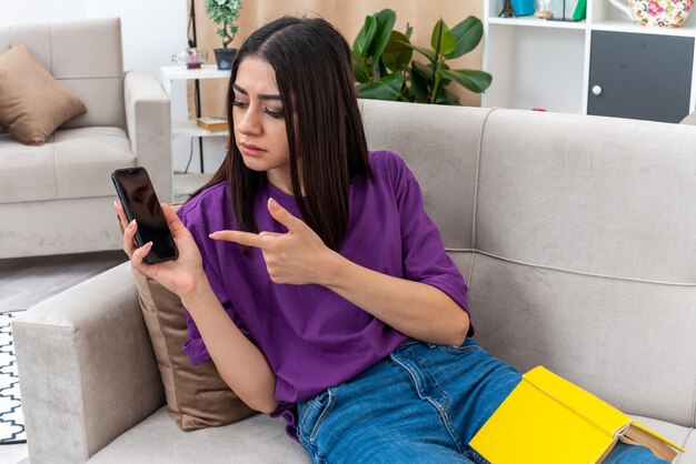 Jeune fille en vêtements décontractés avec livre et smartphone à la confusion assis sur un canapé dans un salon lumineux