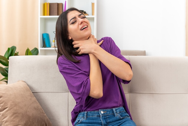 Jeune fille en vêtements décontractés s'étouffant, tenant les mains sur le cou en panique, assise sur un canapé dans un salon lumineux