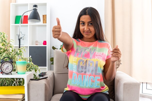 Jeune fille en vêtements décontractés souriant joyeusement montrant les pouces vers le haut assis sur la chaise dans un salon lumineux