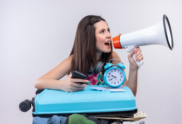 Jeune fille de voyageur caucasien tenant téléphone mobile et haut-parleur avec réveil et valise sur fond blanc isolé