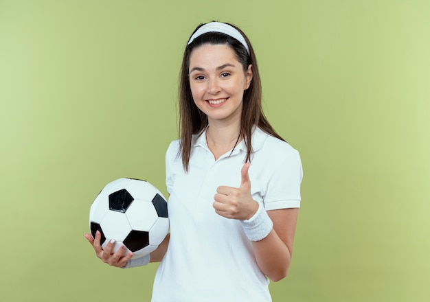 jeune, fitness, femme, dans, bandeau, tenue, ballon football, sourire, projection, pouces haut, debout, sur, mur clair