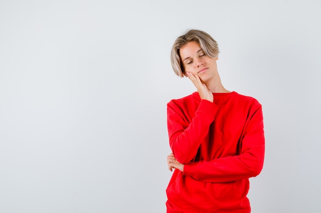 Jeune garçon adolescent appuyé sur la joue en pull rouge et à l'air détendu, vue de face.