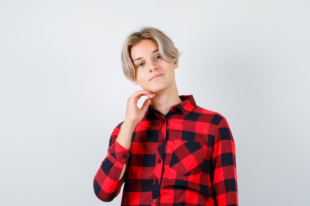 Jeune garçon adolescent gardant la main sous le menton en chemise à carreaux et l'air fier, vue de face.