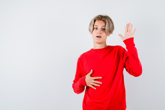 Jeune garçon adolescent montrant la paume, la main sur le ventre en pull rouge et l'air perplexe. vue de face.