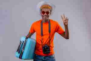 Photo gratuite jeune garçon afro-américain beau voyageur en chapeau d'été portant un t-shirt orange tenant une valise de voyage regardant la caméra souriant sympathique montrant le numéro deux ou signe de la victoire sur backgrou blanc