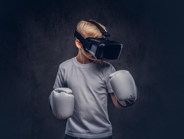 Jeune garçon boxeur aux cheveux blonds vêtu d'un t-shirt blanc portant des lunettes de réalité visuelle et des gants de boxe, entraînement dans un studio. Isolé sur un fond texturé sombre.