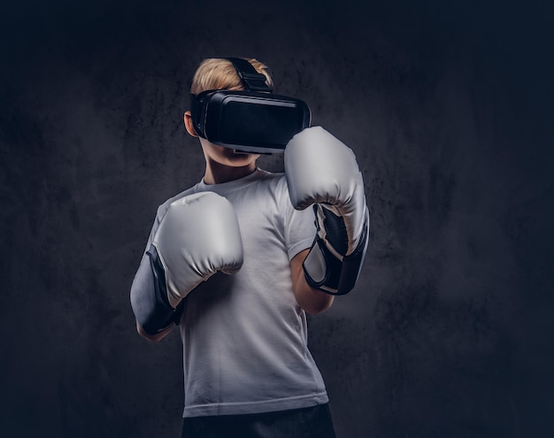 Jeune garçon boxeur aux cheveux blonds vêtu d'un t-shirt blanc portant des lunettes de réalité visuelle et des gants de boxe, entraînement dans un studio. Isolé sur un fond texturé sombre.