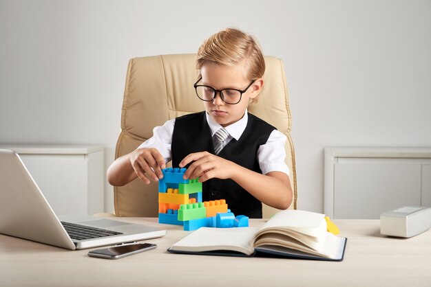 Jeune garçon caucasien blond assis sur une chaise exécutive au bureau et jouant avec des blocs de construction