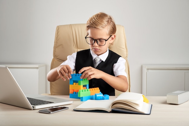 Jeune garçon caucasien blond assis sur une chaise exécutive au bureau et jouant avec des blocs de construction